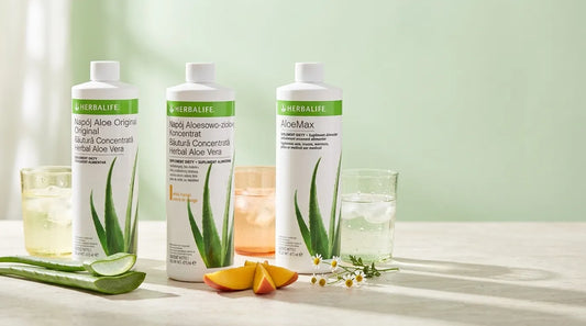 Cele trei băuturi concentrate Aloe Vera Herbalife, Original, Mango și AloeMax, alături de pahare cu apă, frunze de aloe, felii de mango și flori de mușețel